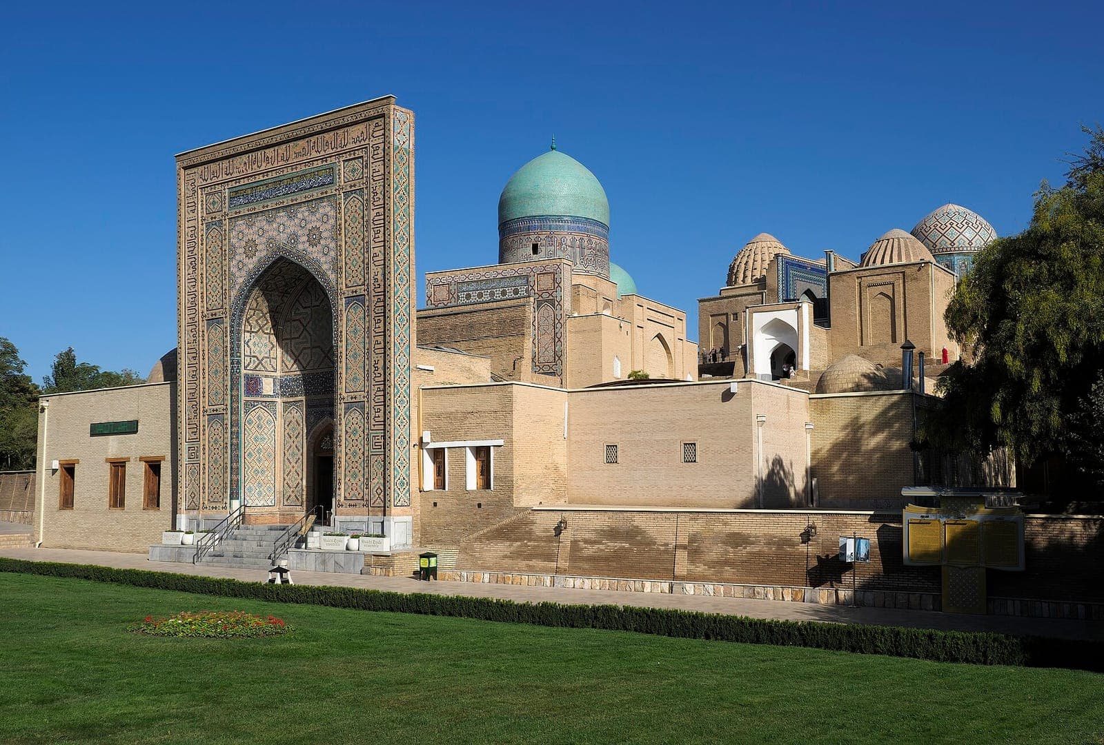 Shah-i-Zinda in Samarkand: a tall tiled portal beside brick domes and a ribbed turquoise cupola, above a lawn.