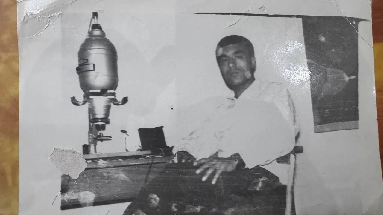 A creased black-and-white photograph of a young man in a white shirt sitting at a photographic enlarger on a wooden bench, one hand resting on the baseboard.