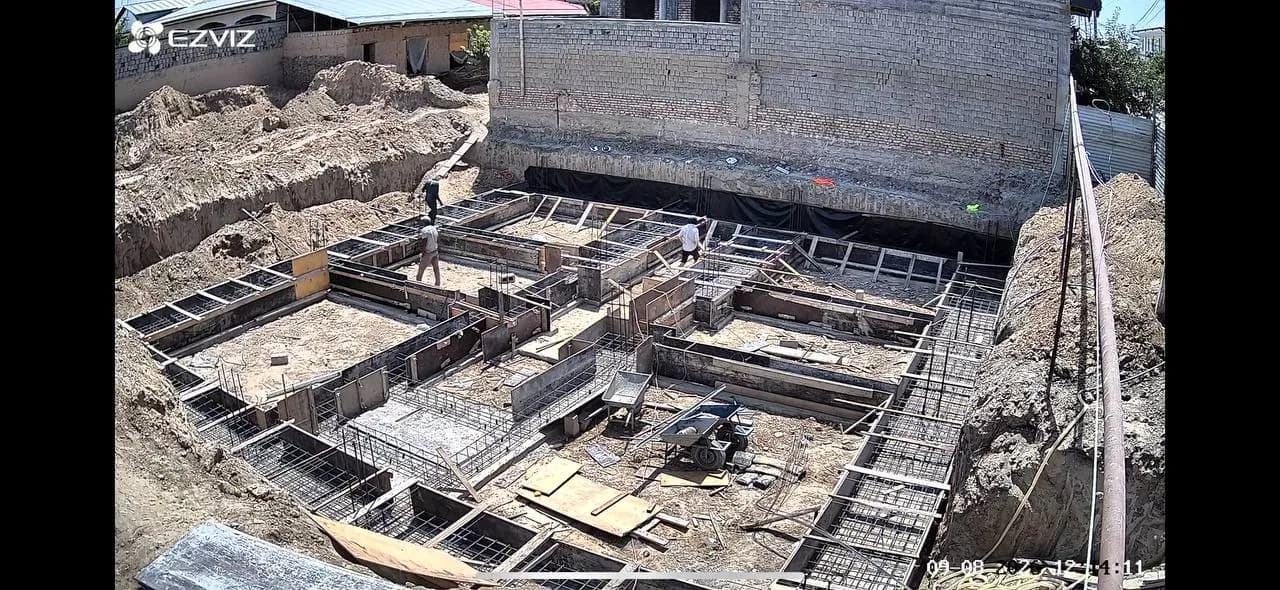 A building site seen from above: concrete foundation trenches laid out as rooms, lined with timber formwork and steel reinforcement, two men walking between them.