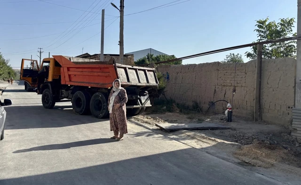 A woman in a patterned dress and white headscarf standing on a newly concreted street beside a tipper truck, a long pakhsa mud-brick wall running behind her.
