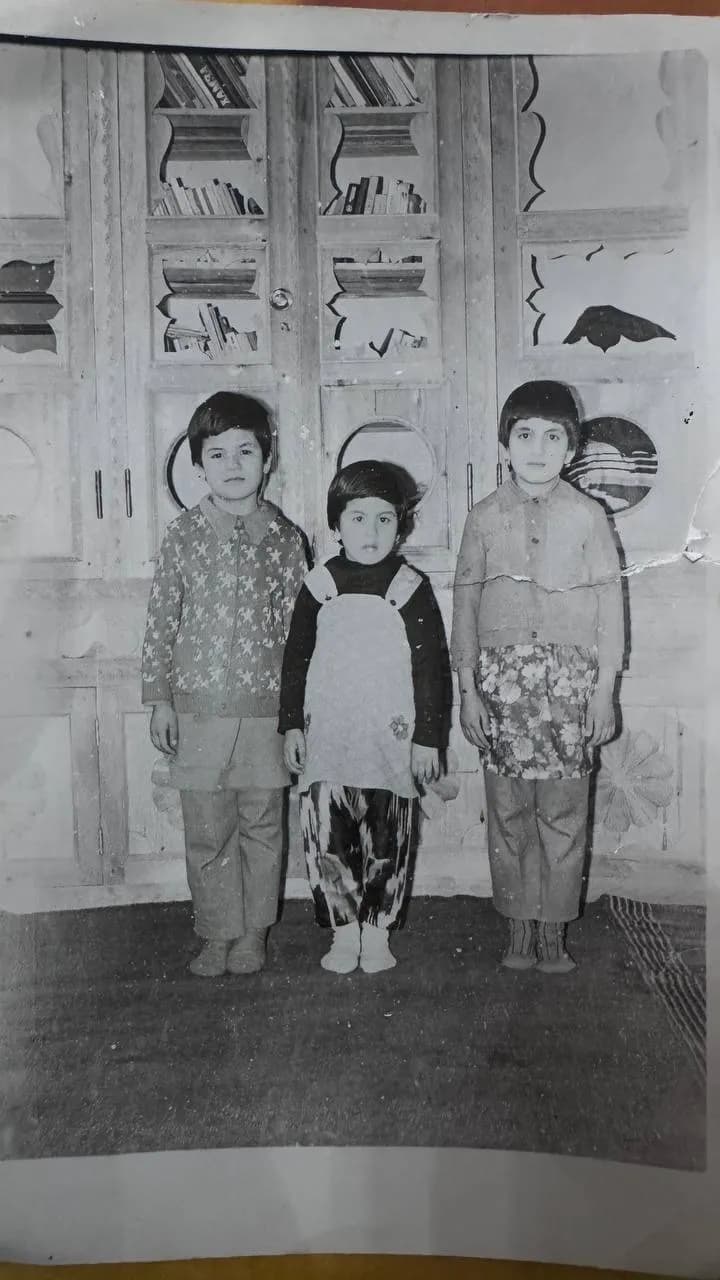 A black-and-white photograph of three children standing on a carpet in front of tall wooden shelves filled with books, the youngest between her two older sisters.